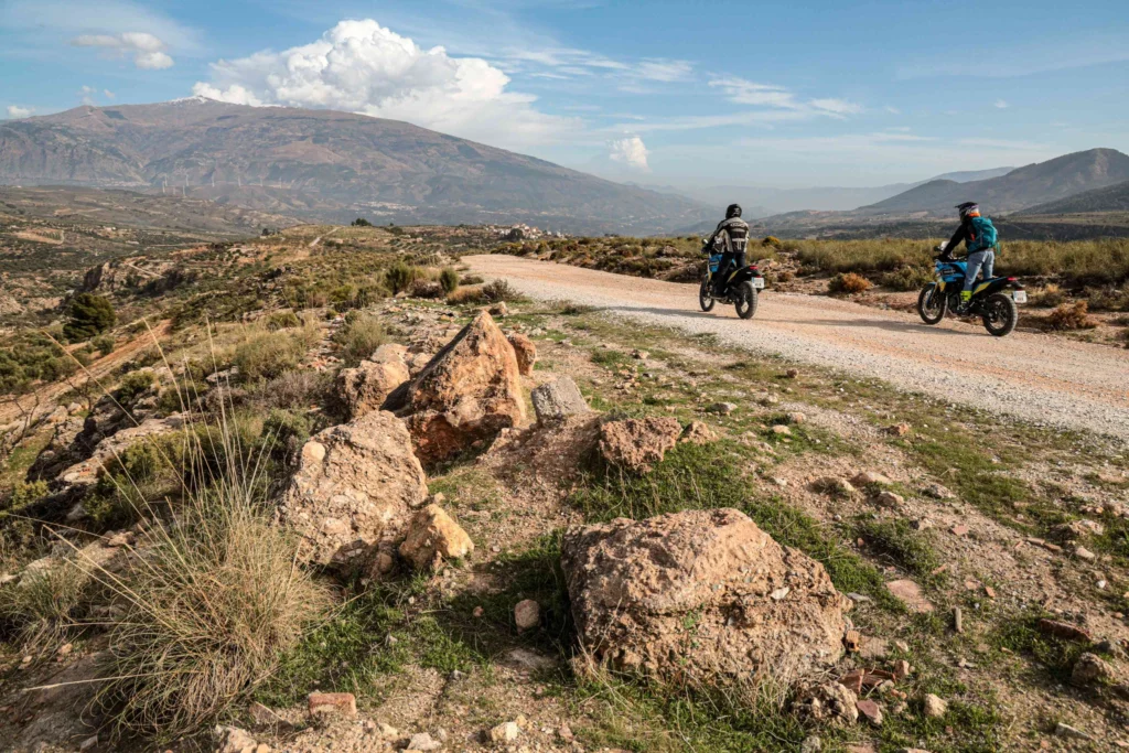 Two adventure motorcyclists riding a wide, accessible gravel trail through the mountains of Andalusia, with a traditional white village in the distance | Dos pilotos de moto trail recorriendo una pista de grava ancha y accesible por las montañas de Andalucía, con un pueblo blanco tradicional a lo lejos.