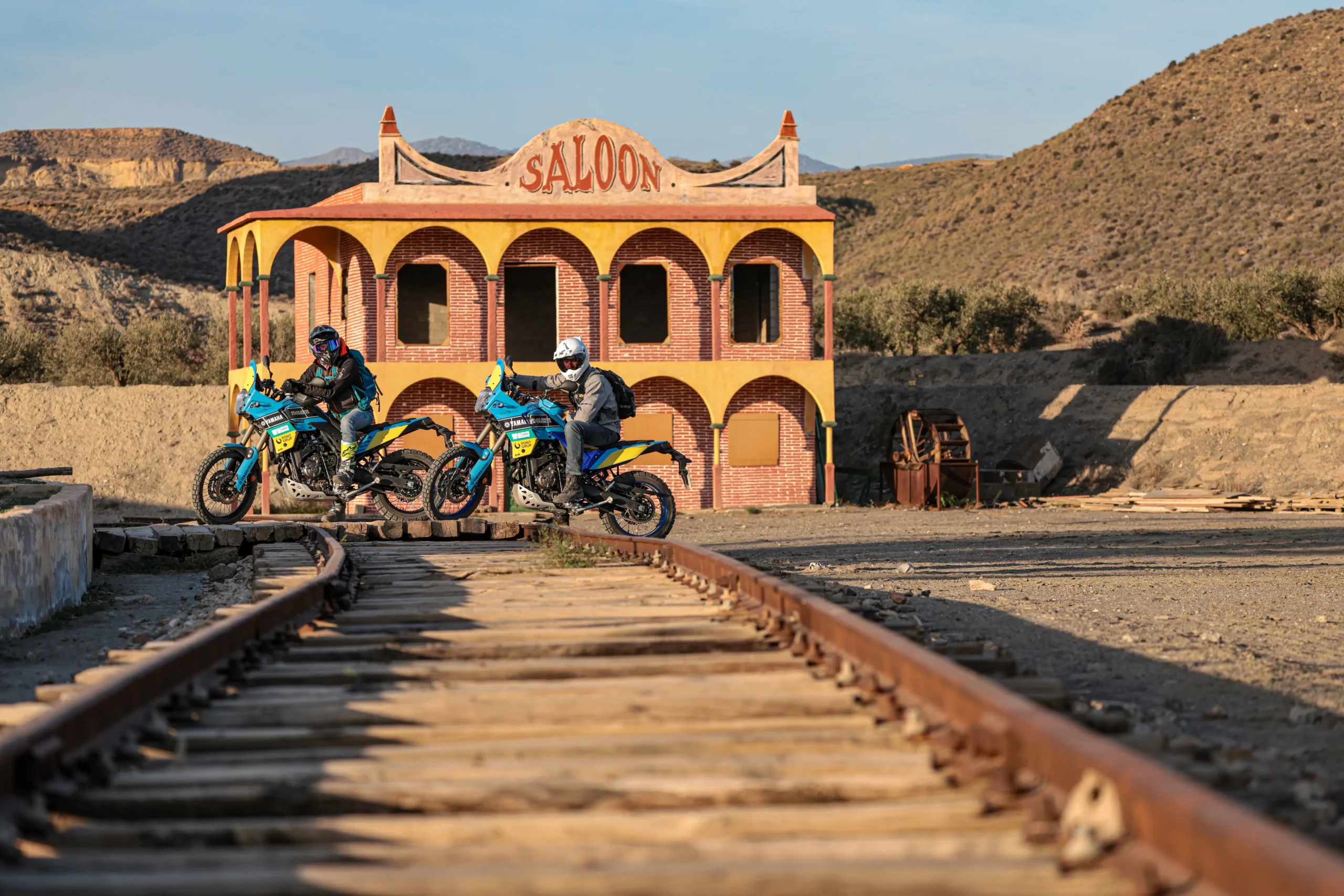 Two adventure motorcycle riders crossing train tracks in front of a western saloon in the Tabernas Desert, Almeria | Dos motoristas de aventura cruzando las vías del tren frente a un poblado del oeste en el desierto de Tabernas, Almería.