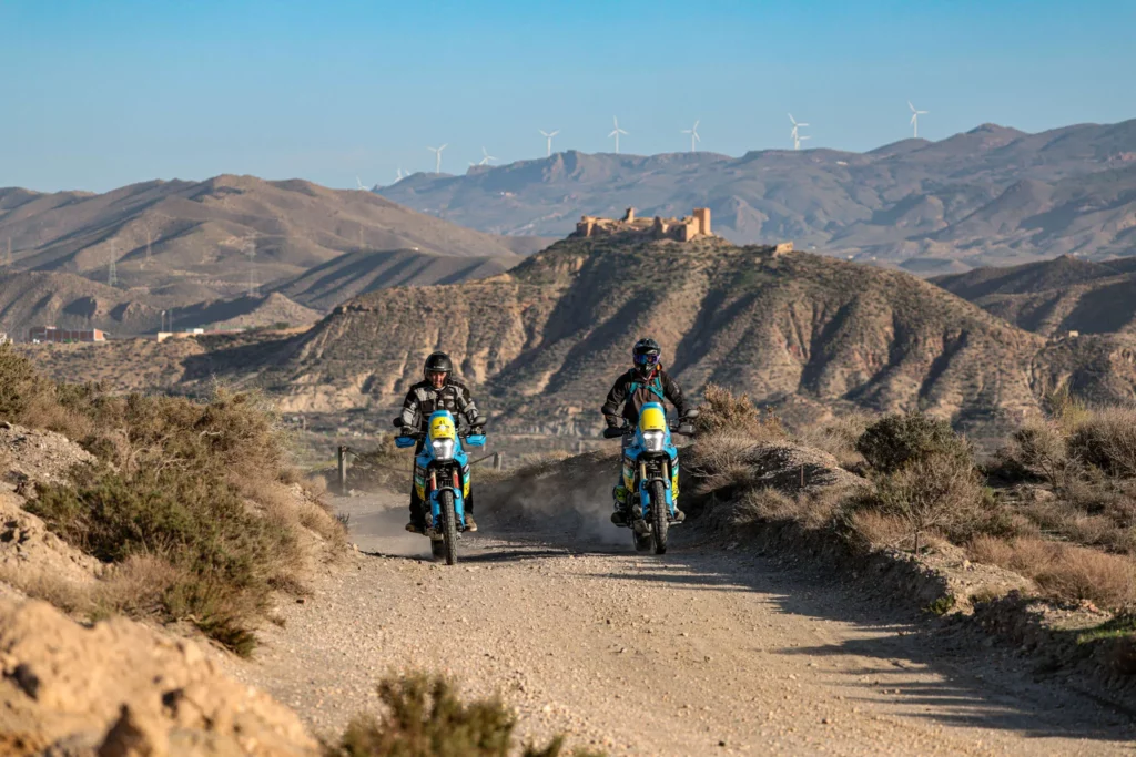 Two adventure motorcyclists enjoying a sunny, accessible dirt track in Andalusia, with historic castle ruins in the background | Dos pilotos de moto trail disfrutando de una pista de tierra soleada y accesible en Andalucía, con las ruinas de un castillo histórico al fondo.