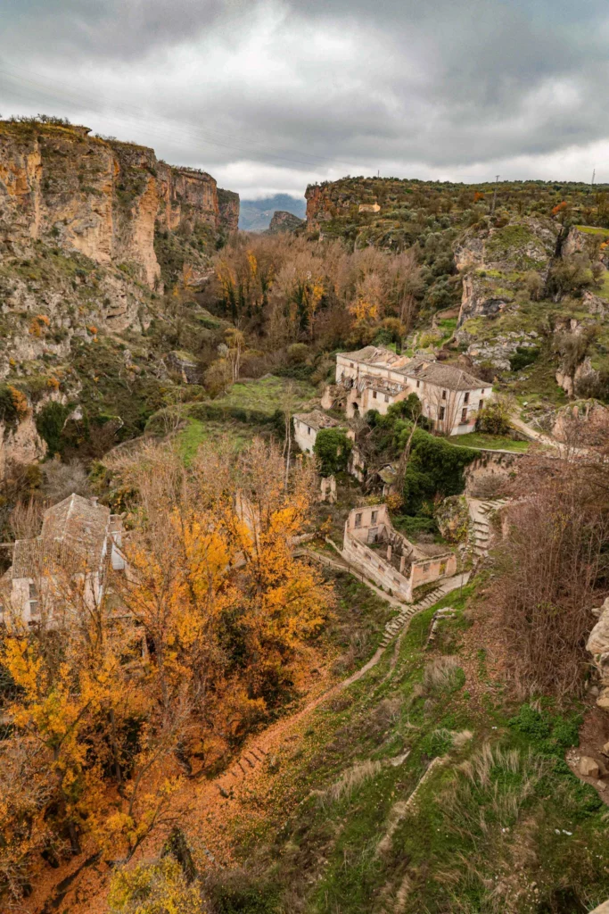 A deep mountain gorge featuring abandoned stone buildings and autumn trees, highlighting the hidden off-road motorcycle routes available in Andalusia, Spain | Un profundo desfiladero montañoso con edificios de piedra abandonados y árboles otoñales, destacando las rutas ocultas de moto trail disponibles en Andalucía, España.