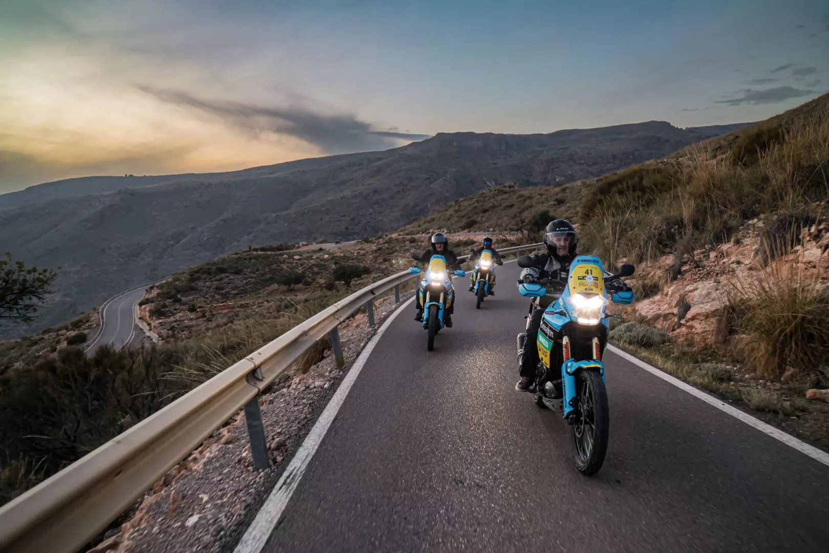 A group of adventure motorcyclists riding on a winding paved mountain road at sunset during a guided tour in Andalusia, Spain. | Un grupo de motociclistas de aventura conduciendo por una sinuosa carretera de montaña asfaltada al atardecer durante una ruta guiada en Andalucía, España.