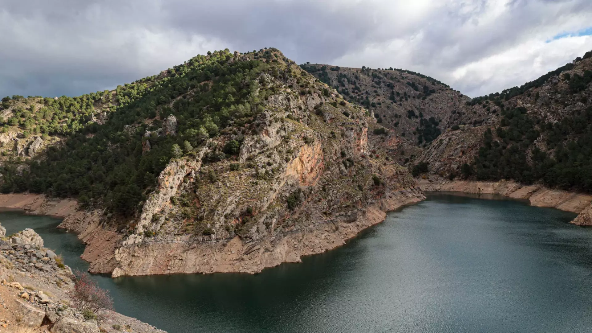 A spectacular view of a deep blue mountain reservoir surrounded by rugged rocky cliffs and pine trees, a scenic highlight on our Andalusia motorcycle tours. | Una vista espectacular de un embalse de montaña azul profundo rodeado de acantilados rocosos y pinos, un paisaje destacado en nuestras rutas en moto por Andalucía.