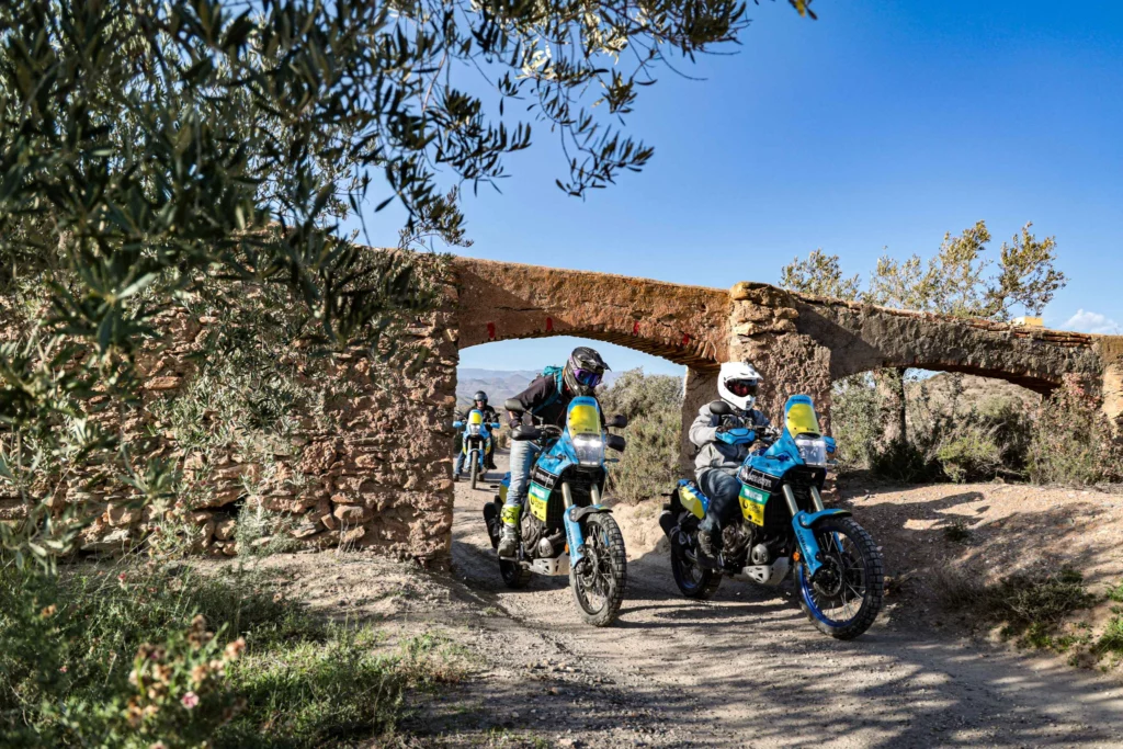 A group of adventure motorcycle riders navigating a dirt trail under a historic stone archway during a guided off-road tour in Andalusia, Spain. | Un grupo de pilotos de motos trail navegando por un camino de tierra bajo un arco de piedra histórico durante una ruta guiada off-road en Andalucía, España.