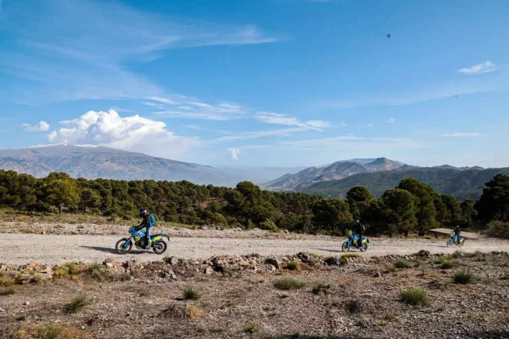 A group of three adventure motorcyclists riding on a wide, high-altitude dirt trail with the snow-capped Sierra Nevada mountains in the background during a tour in Andalusia, Spain. | Un grupo de tres motociclistas de aventura conduciendo por una pista de tierra a gran altitud con las montañas nevadas de Sierra Nevada al fondo durante una ruta en Andalucía, España.