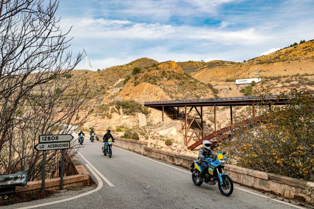 A group of adventure motorcycle riders crossing a bridge on a winding mountain road near Izbor, showcasing the scenic connecting routes of our Andalusian motorcycle tours. | Un grupo de motociclistas de aventura cruzando un puente en una sinuosa carretera de montaña cerca de Ízbor, mostrando las pintorescas rutas de conexión de nuestros tours en moto por Andalucía.